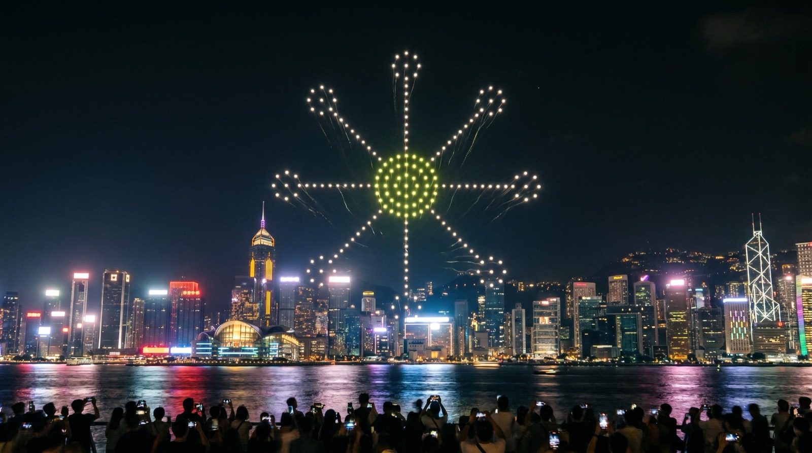 A drone light show forming the IAW hub mark over a harbour skyline — brand visual